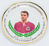 Farbig bedruckter Wandteller mit dem Portrt von Karl Mai und der Inschrift "23facher Deutscher Nationalspieler. Karl Mai Weltmeister 1954 Endspiel gegen Ungarn in Bern"  mit faksimilierter Unterschrift von Mai. Porzellan, ca. 1974, 19 cm.