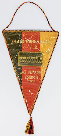 Souvenirseidenwimpel mit aufgesticktem Schriftzug England - Deutschland 4:2. WM-Endspiel London 1966 mit aufgesetztem Mannschaftsfoto des deutschen Teams umrankt von Goldbrokatband. Mit schwarz-gelber Seidenbordre. 40x28 cm (ohne Bommel).<br>-- Schtzpr